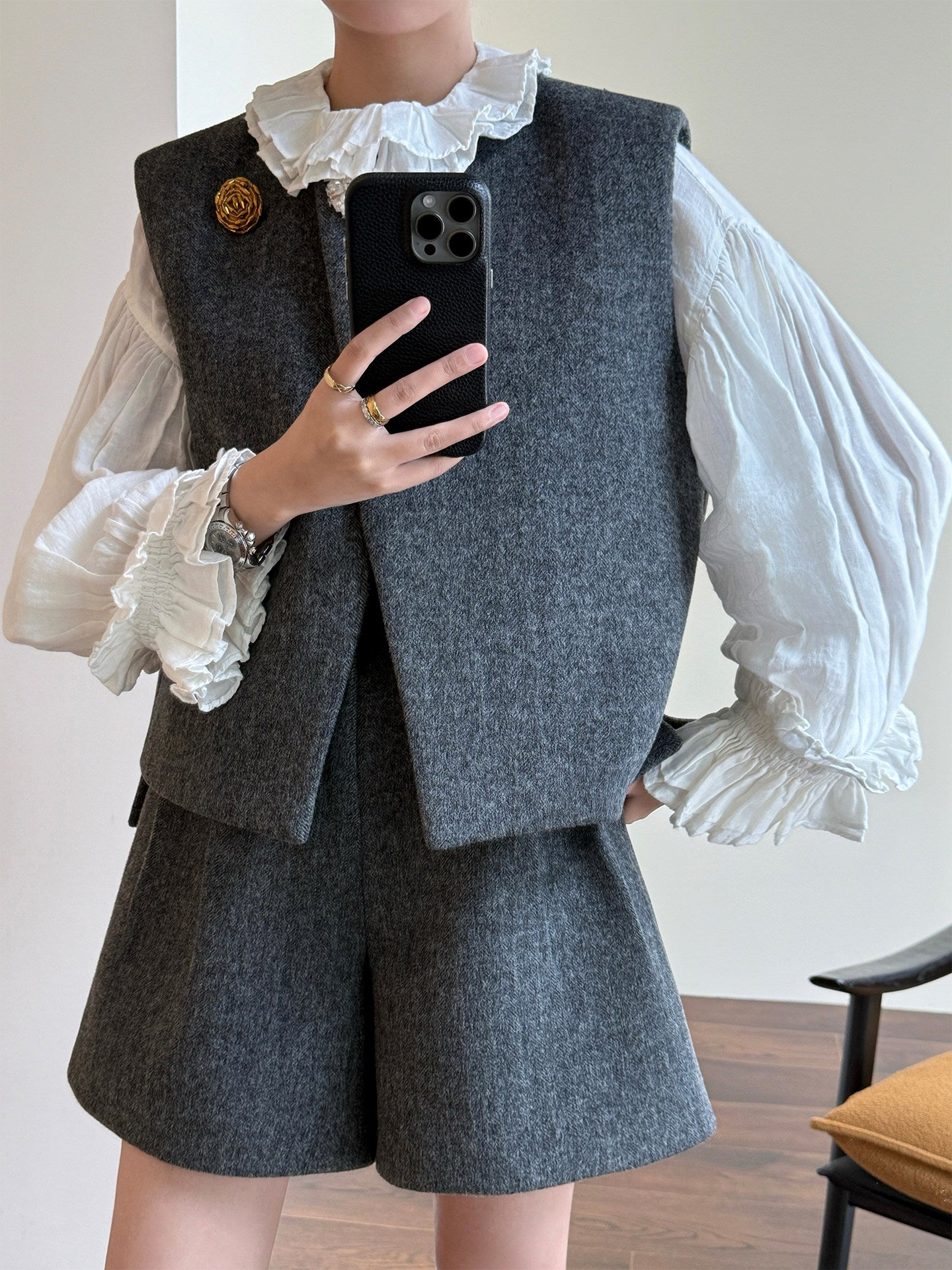 A person takes a mirror selfie showing a gray two-piece vest and shorts set with a white ruffled blouse. The vest has a gold decorative pin on the chest.