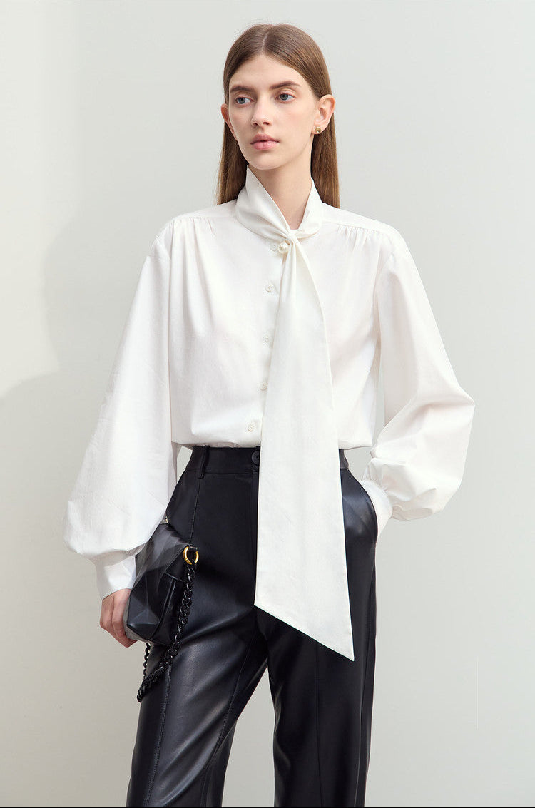 A woman modeling a white long-sleeve blouse with a large bow tie-neck detail, paired with black pants and a black shoulder bag, against a plain light gray studio background.