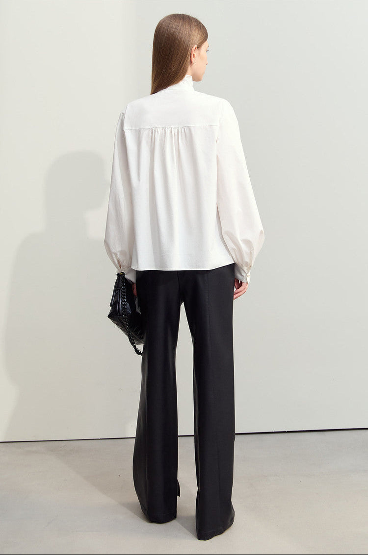A model shown from the back wearing a white long-sleeve blouse with a gathered neckline and black wide-leg trousers, holding a small black textured handbag against a minimalist studio background.