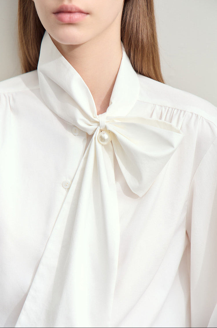 A close-up of a model wearing a white long-sleeve blouse with a large bow tie detail at the collar and a pearl accent.