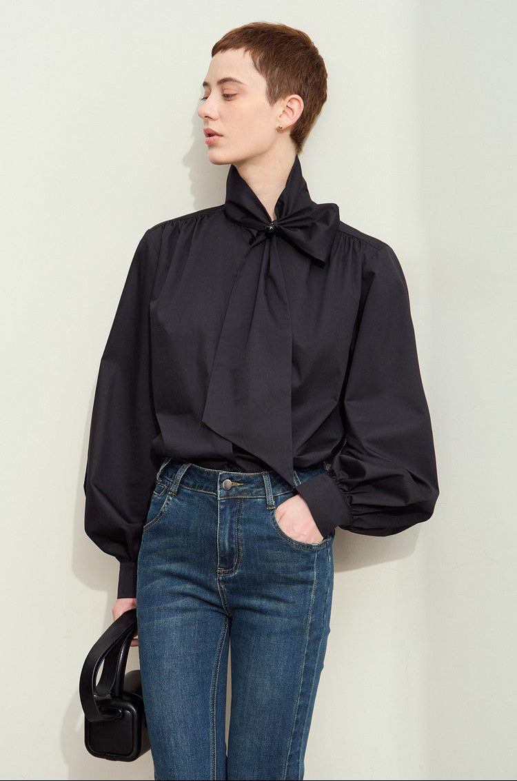A model wearing a black long-sleeve blouse with a large bow at the collar and blue jeans, standing against a light gray wall.