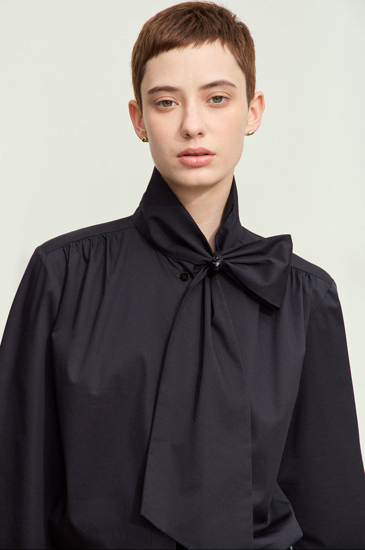 A model with short brown hair wearing a black long-sleeve blouse with a high gathered collar and a large bow detail at the neck, photographed against a light gray studio background.