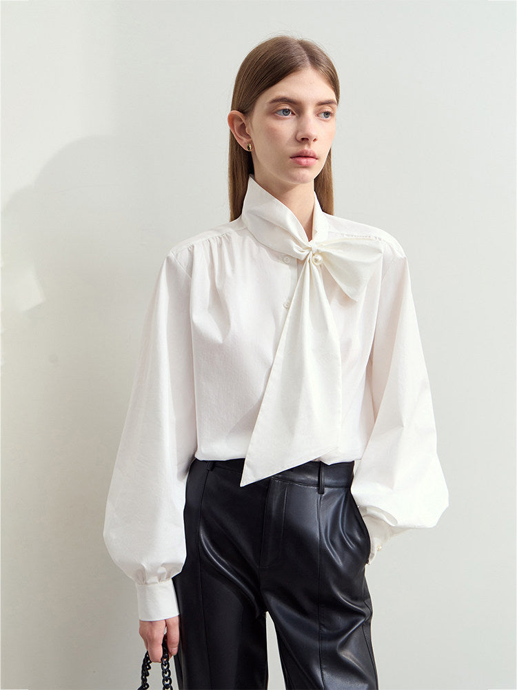 A woman modeling a white long-sleeve blouse with a large bow at the collar and black leather pants, standing against a plain light gray wall.