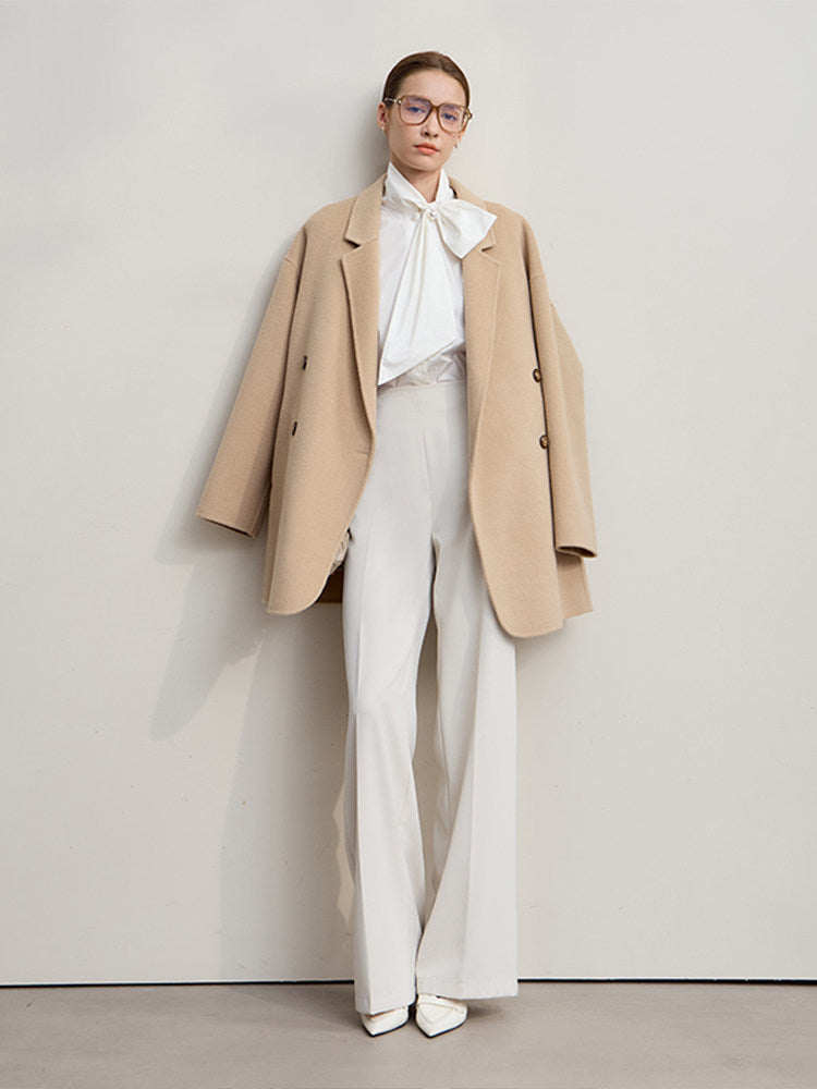 A model stands against a plain light gray wall wearing a beige double-breasted blazer over a white high-neck bow blouse, white wide-leg trousers, and white pointed shoes.