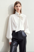 A woman models a white long-sleeve blouse with a large bow at the collar and black high-waisted pants, standing against a light wall. The top of the image has a white band with Chinese text '未伖色'.