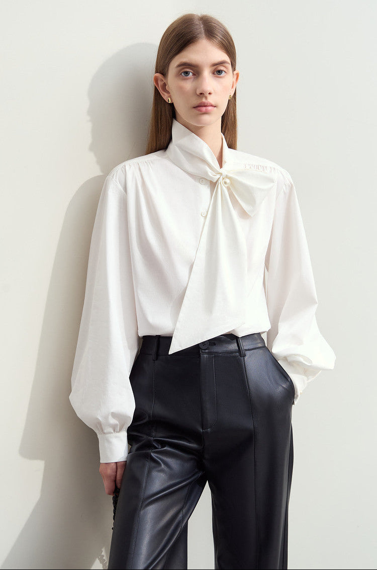 A woman models a white long-sleeve blouse with a large bow at the collar and black high-waisted pants, standing against a light wall. The top of the image has a white band with Chinese text '未伖色'.