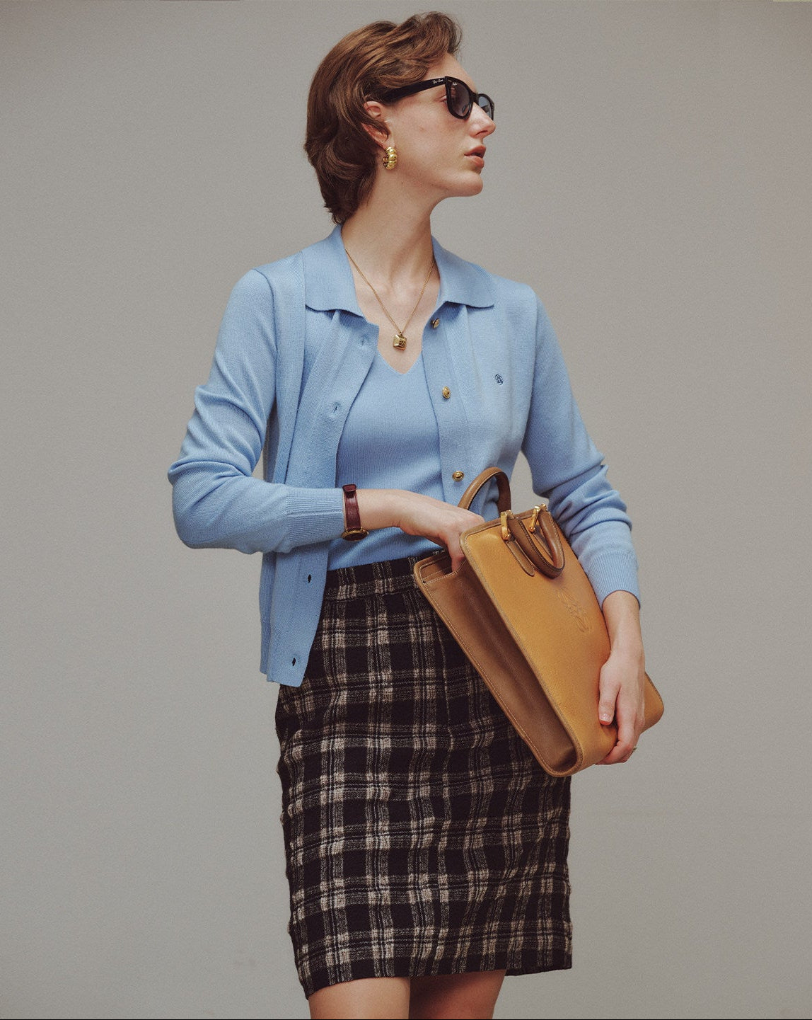 Woman in a light blue jacket and plaid skirt holding a tan handbag against a plain background