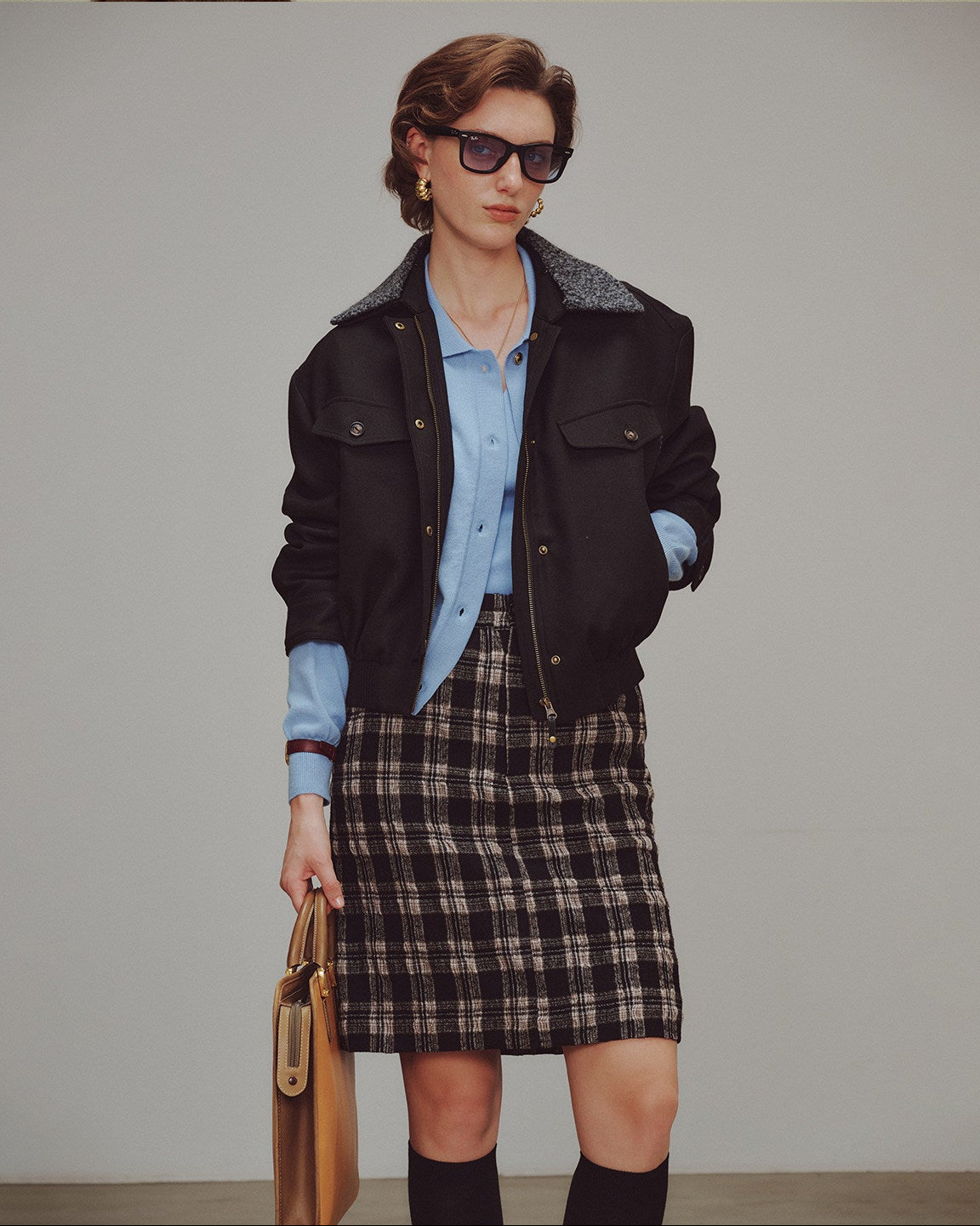 Woman wearing a black jacket, blue shirt, plaid skirt, and sunglasses against a plain background