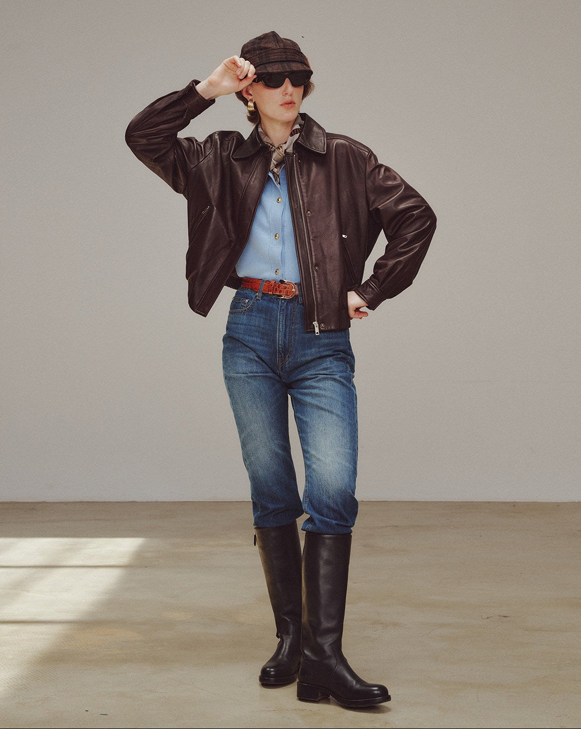 woman wearing a brown leather jacket, blue jeans, and black boots in a minimalistic setting.