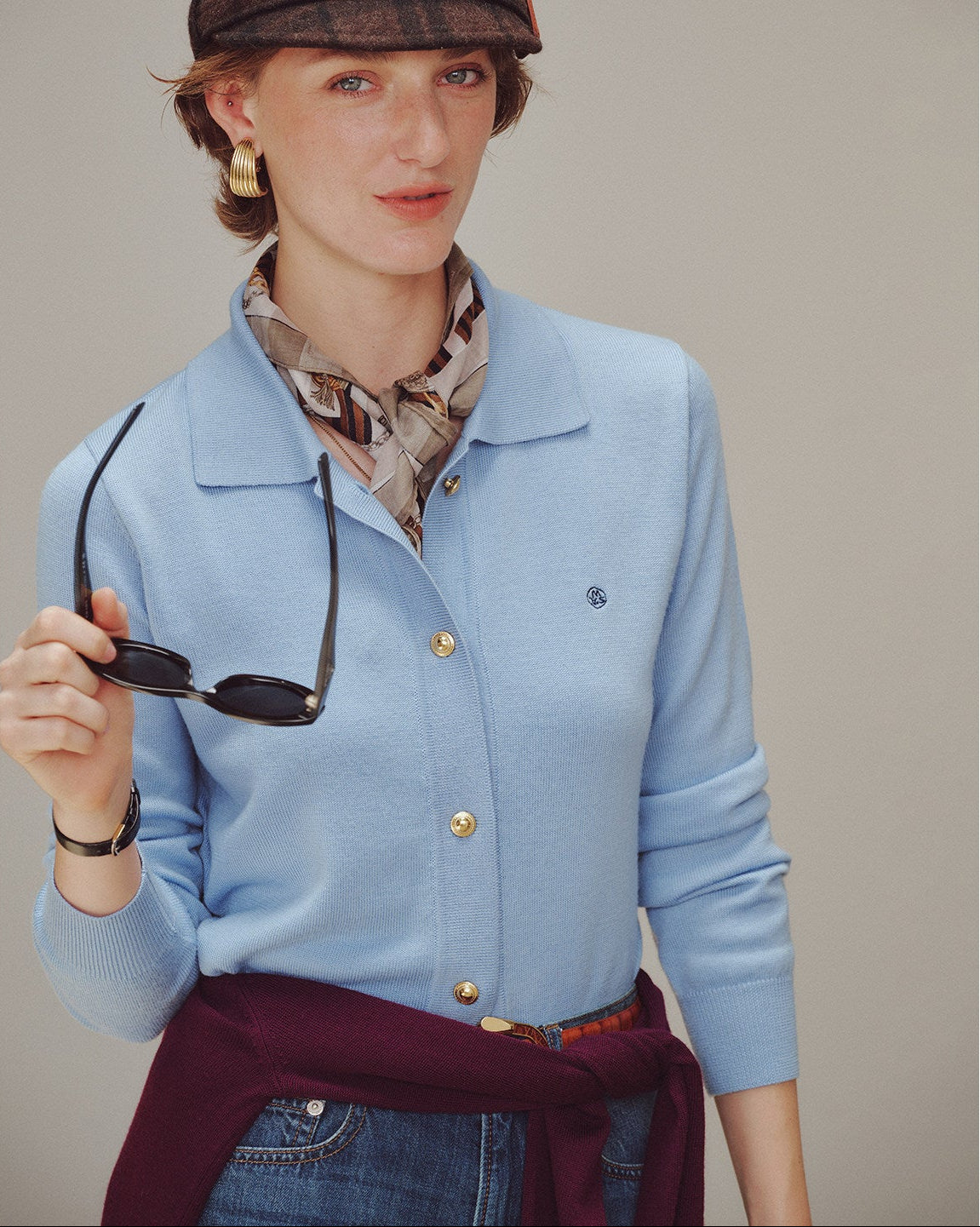 Woman wearing a light blue cardigan with gold buttons, holding sunglasses, against a plain background.