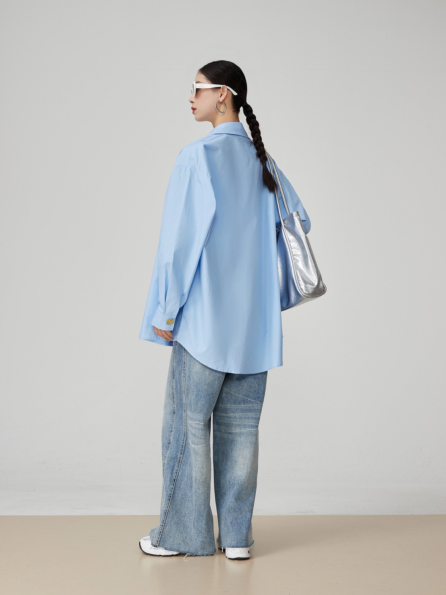 woman wearing a light blue oversized shirt and jeans, holding a silver bag against a plain background