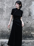 A woman standing against a textured wall, wearing a long, black, velvet cheongsam dress with a crew neckline and three-quarter sleeves.