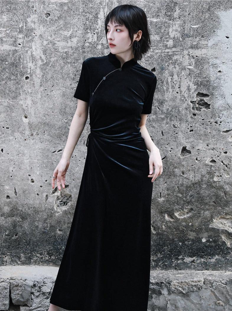 A woman standing against a textured wall, wearing a long, black, velvet cheongsam dress with a crew neckline and three-quarter sleeves.