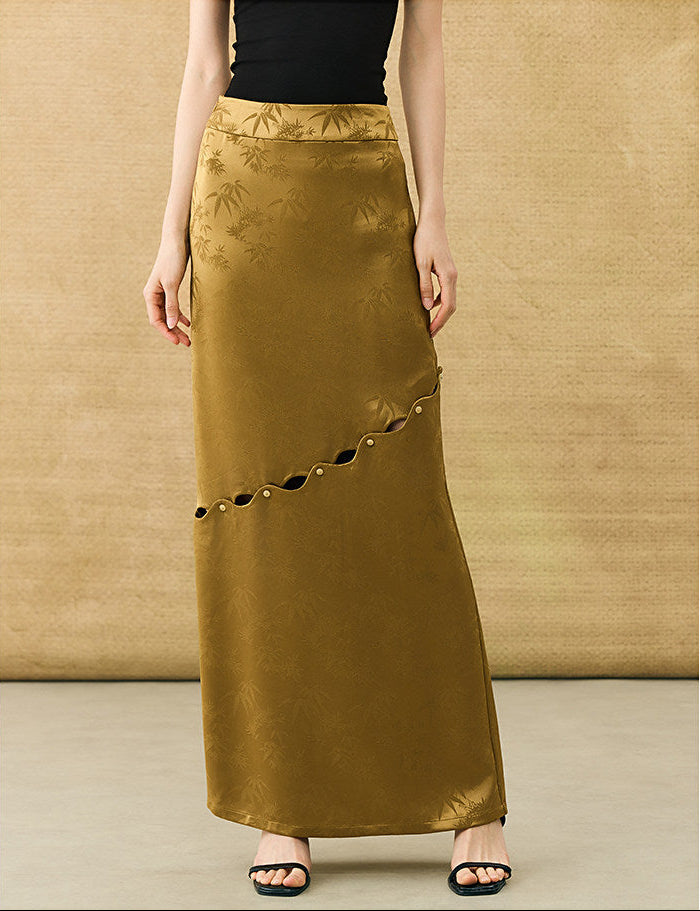 A woman models a long, floor-length olive-gold skirt with a subtle tonal bamboo pattern, decorative side cutouts with gold-tone rings, and a black top. The background is a textured beige wall and floor.