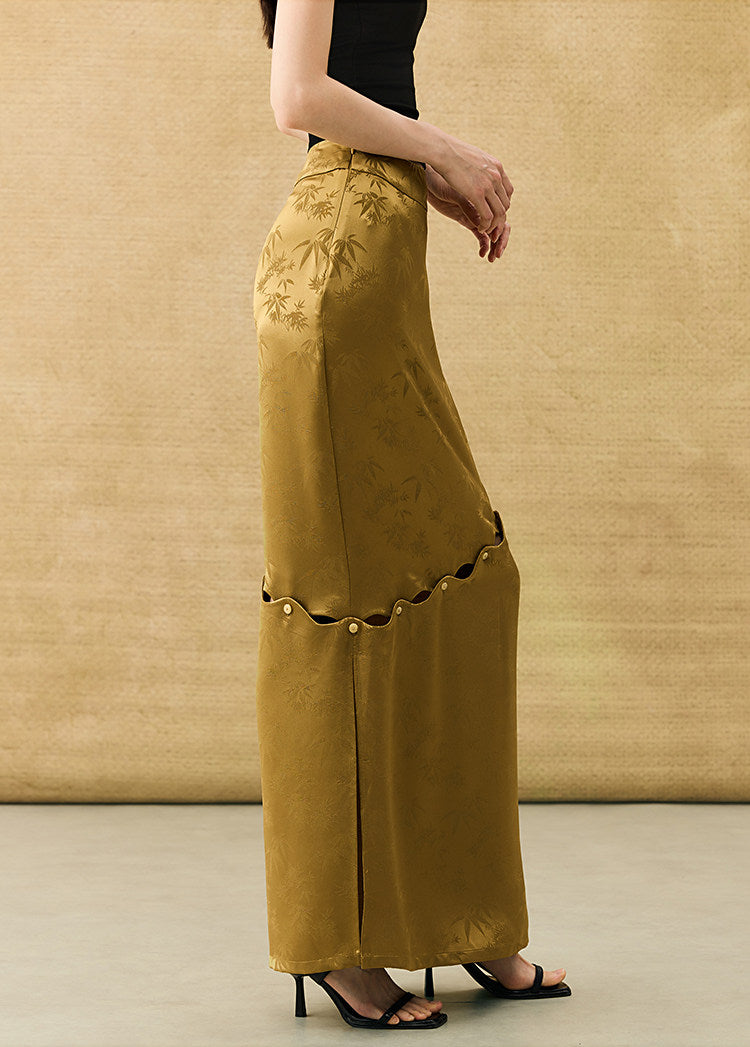 A model is shown from the side wearing a mustard yellow wide-leg skirt with tonal floral embroidery and decorative cutout details, paired with a black top and black sandals, set against a textured beige backdrop.