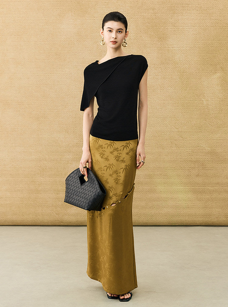 A model stands against a textured beige backdrop wearing a black asymmetrical short-sleeve top and a gold maxi skirt with tonal floral embroidery. She holds a black quilted handbag.