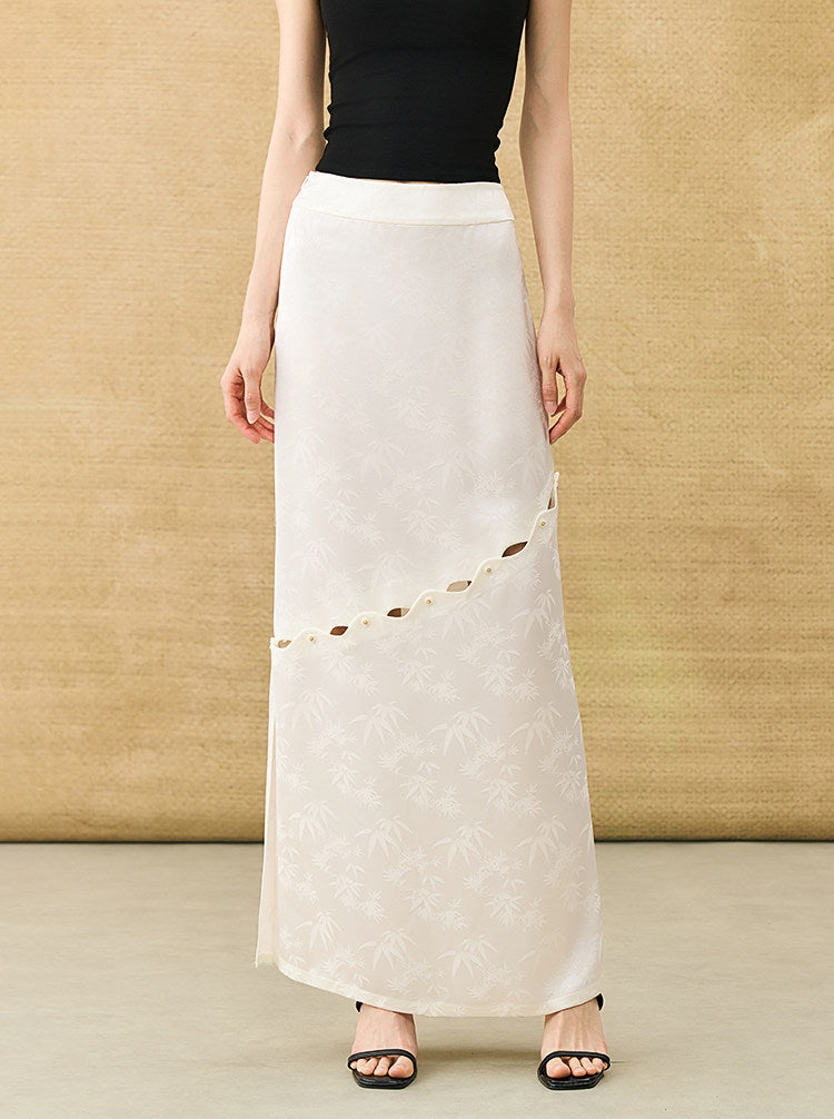 A model wearing a long, flowing off-white maxi skirt with a subtle tonal floral pattern and a diagonal cutout detail, styled with a black top and black sandals against a textured beige backdrop.
