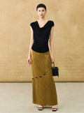 A woman stands against a textured beige backdrop wearing a black draped sleeveless top and a long mustard-yellow skirt with a diagonal cutout detail. She is holding a small black beaded handbag and wearing black sandals.