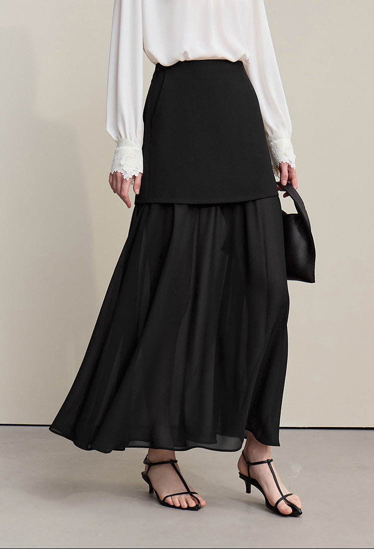 A model wearing a black high-waisted, layered maxi skirt paired with a white blouse, black strappy heels, and holding a black handbag against a neutral beige studio background.
