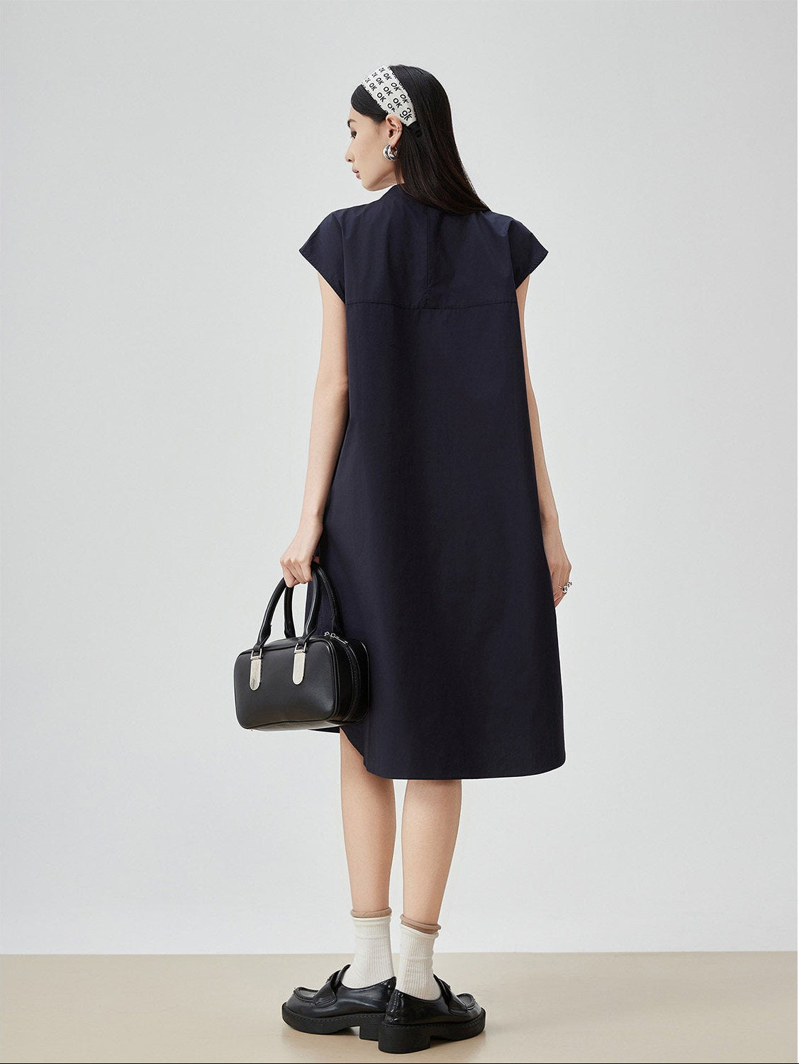 Woman wearing a black dress holding a black handbag against a plain background