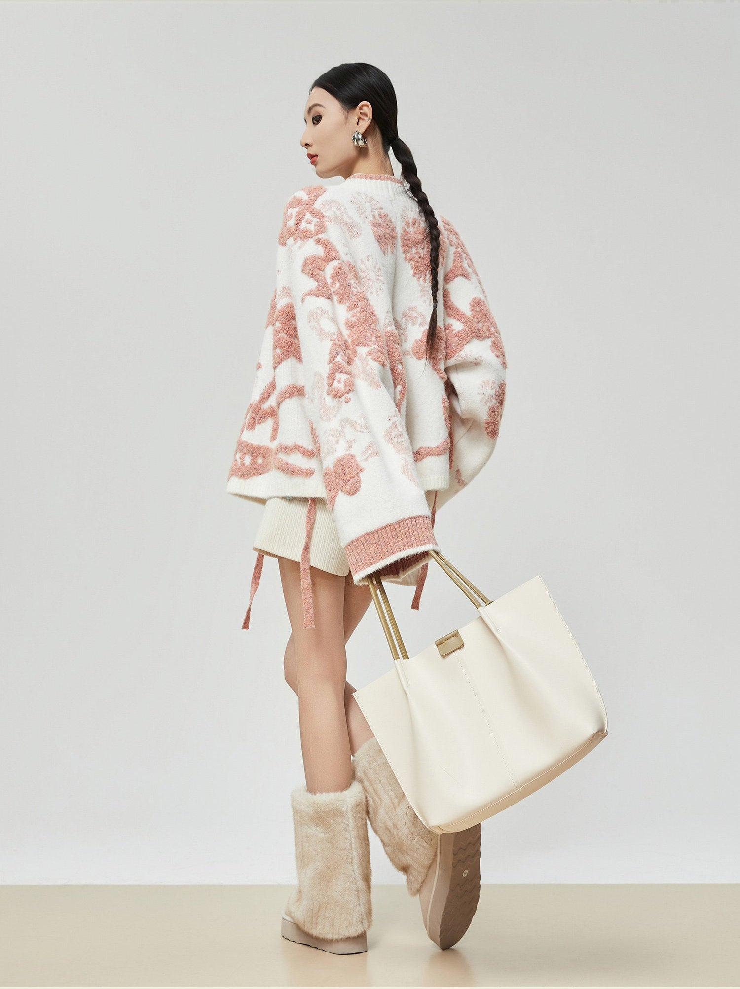 Woman wearing a floral patterned coat, holding a cream-colored bag, and standing against a plain background.