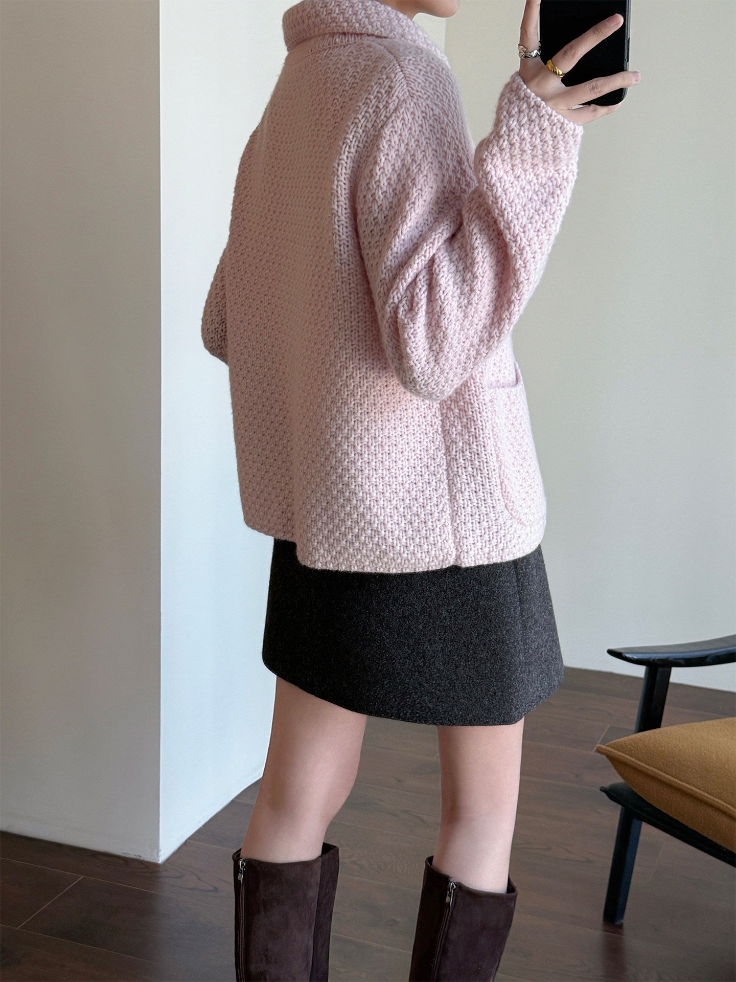 A person wearing a light pink textured knit cardigan, a dark gray mini skirt, and dark brown boots, taking a mirror selfie in an indoor setting.