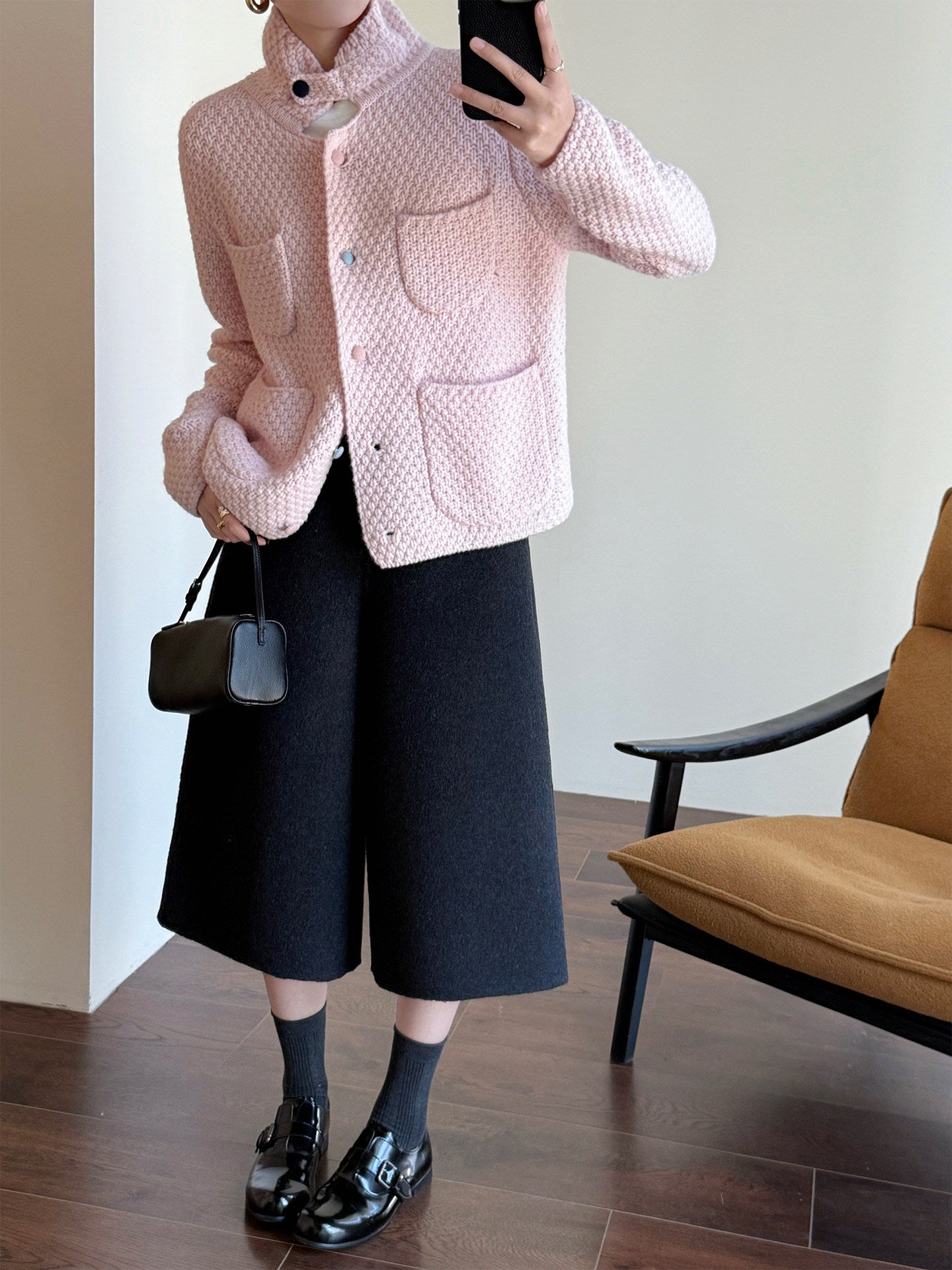 A person takes a mirror selfie indoors wearing a light pink textured jacket, black wide-leg cropped pants, black socks, and black patent leather loafers, while holding a small black handbag.