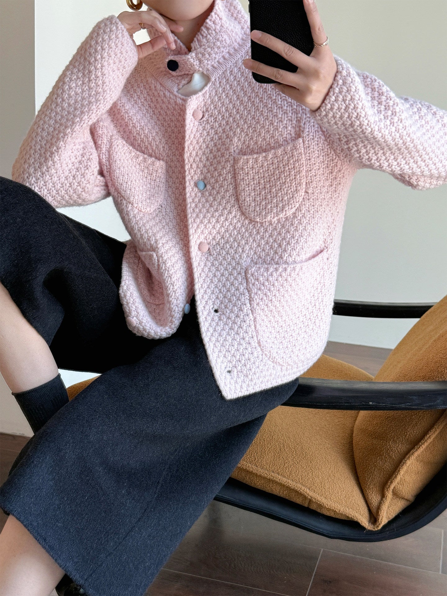 A person seated indoors wearing a light pink textured jacket with large front patch pockets and dark charcoal skirt or pants, holding a phone to take a mirror selfie. A mustard chair is visible in the background.