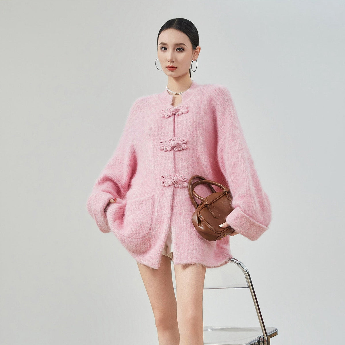 A woman models a pink fuzzy cropped cardigan with decorative frog closures, paired with a brown handbag, against a plain light gray studio background.