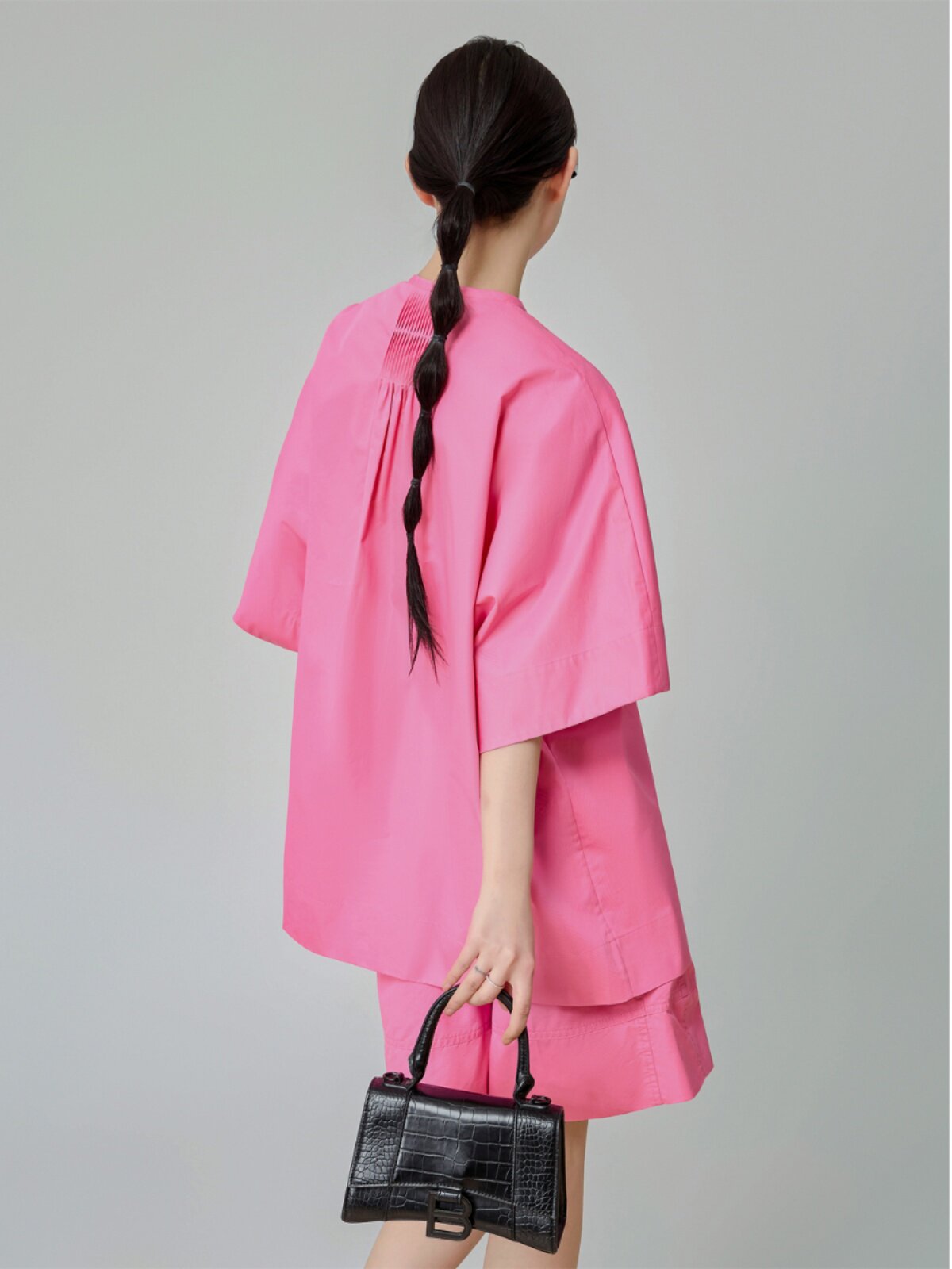 Person wearing a bright pink outfit holding a black handbag on a gray background