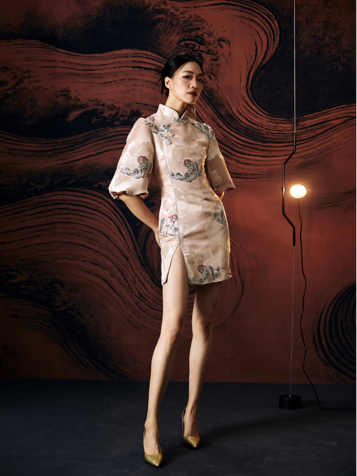 A woman standing against a textured wall, wearing a short, pink, jacquard floral cheongsam dress with a stand collar neckline and three-quarter sleeves
