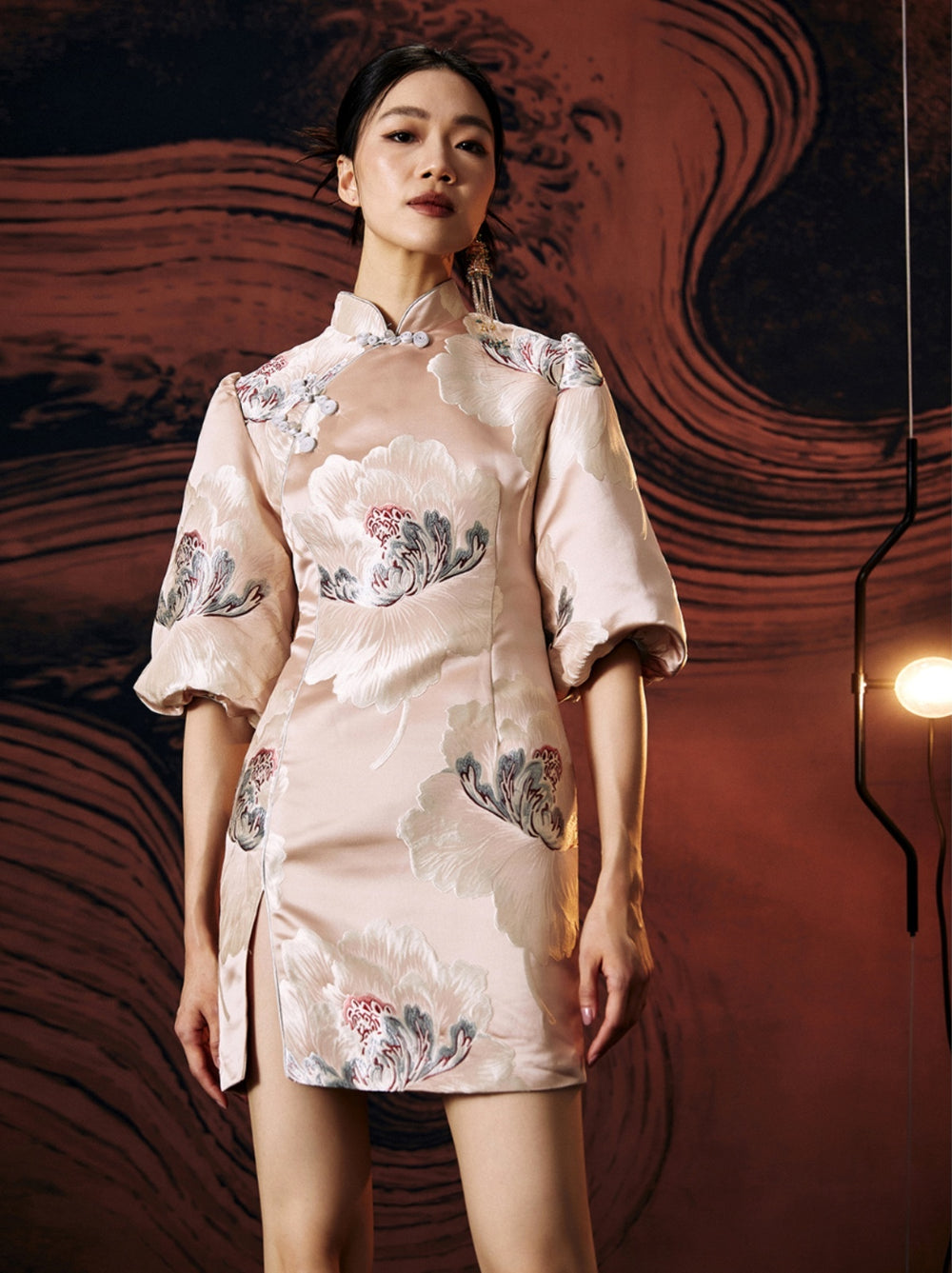 A woman standing against a textured wall, wearing a short, pink, jacquard floral cheongsam dress with a stand collar neckline and three-quarter sleeves
