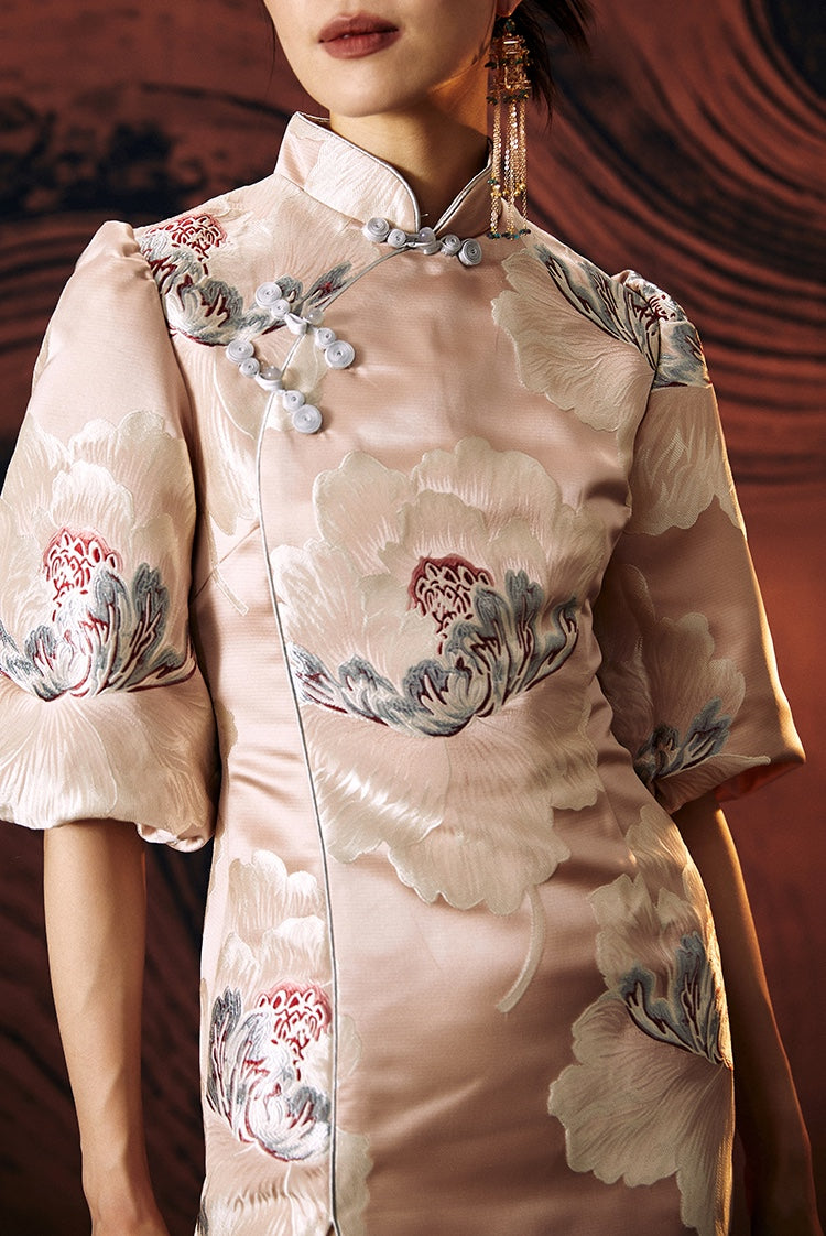 A woman standing against a textured wall, wearing a short, pink, jacquard floral cheongsam dress with a stand collar neckline and three-quarter sleeves