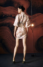 A woman standing against a textured wall, wearing a short, pink, jacquard floral cheongsam dress with a stand collar neckline and three-quarter sleeves