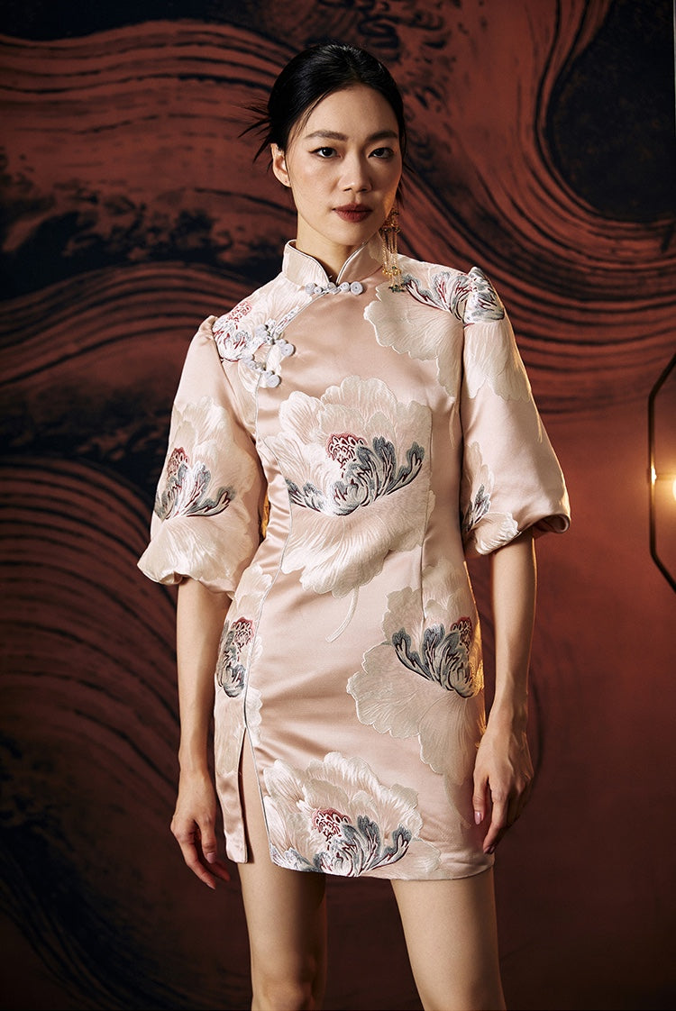A woman standing against a textured wall, wearing a short, pink, jacquard floral cheongsam dress with a stand collar neckline and three-quarter sleeves