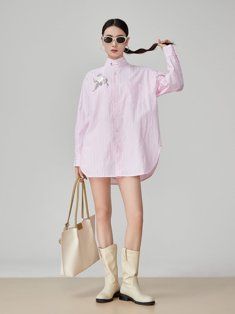 Woman wearing a pink striped shirt, beige knee-high boots, and holding a beige handbag against a gray background