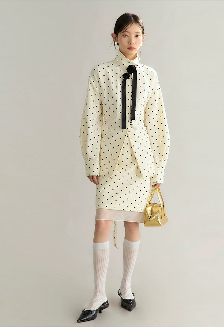 Woman wearing a polka dot dress with a high collar and a black bow, holding a gold handbag on a plain background.