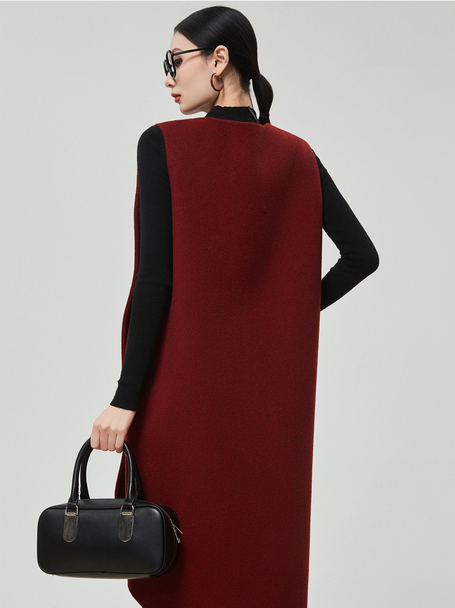 Woman wearing a long red vest over a black top, holding a black handbag on a light gray background