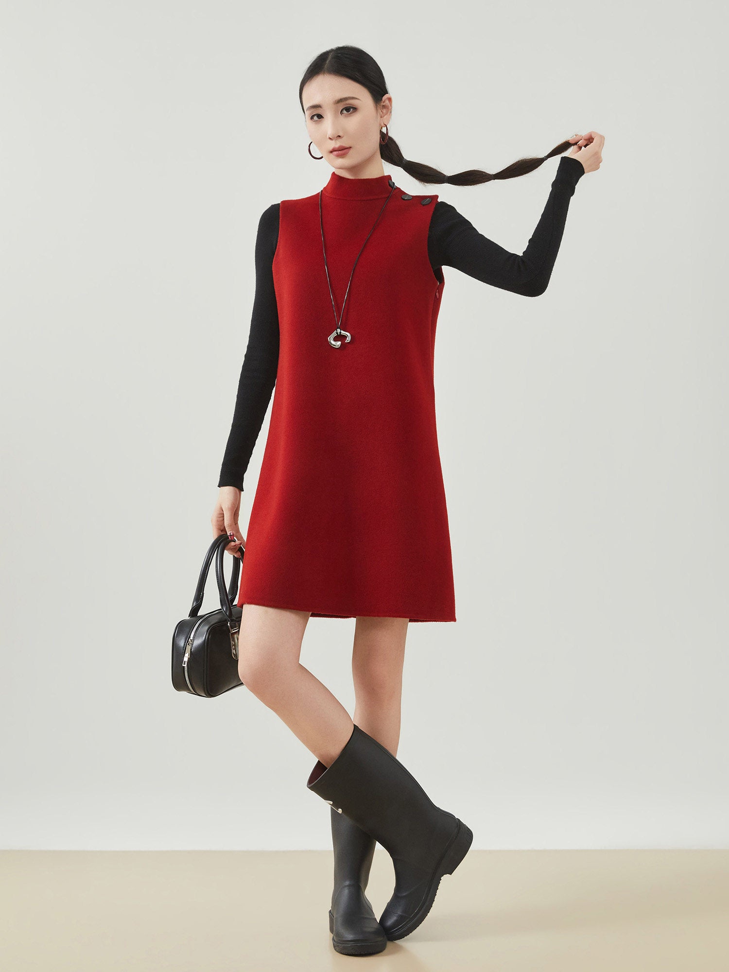 Woman wearing a red sleeveless dress with a black long-sleeve top underneath, holding a black handbag, and wearing black rain boots on a white background.