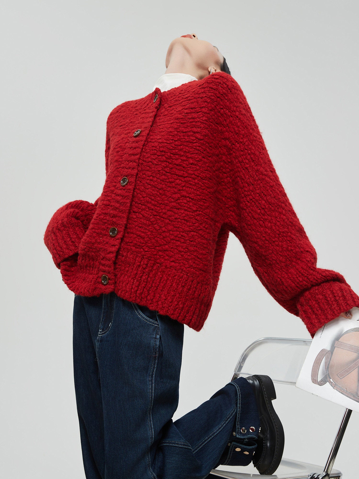 Woman wearing a red knitted cardigan and dark jeans sitting on a white chair against a light gray background