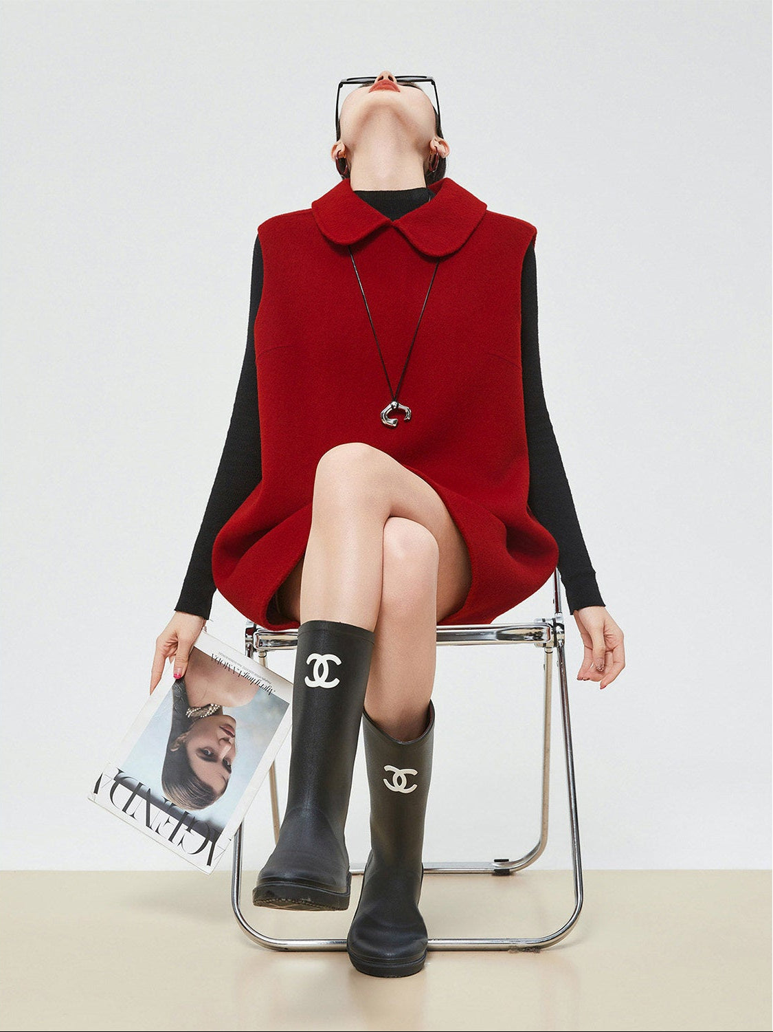 Person wearing a red vest over a black dress with black rain boots, sitting on a chair.