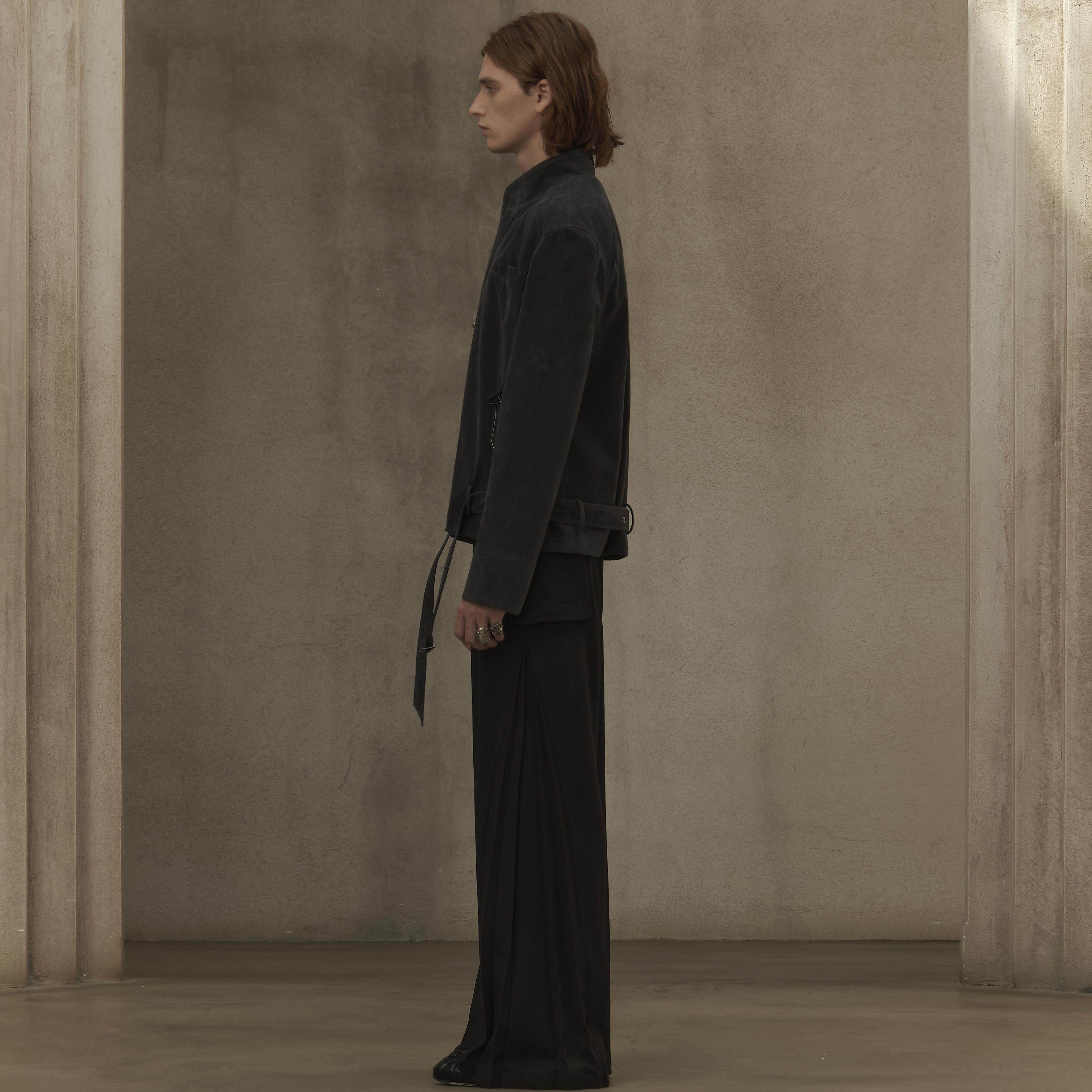 Person wearing a dark outfit standing in a minimalistic room with a neutral wall.