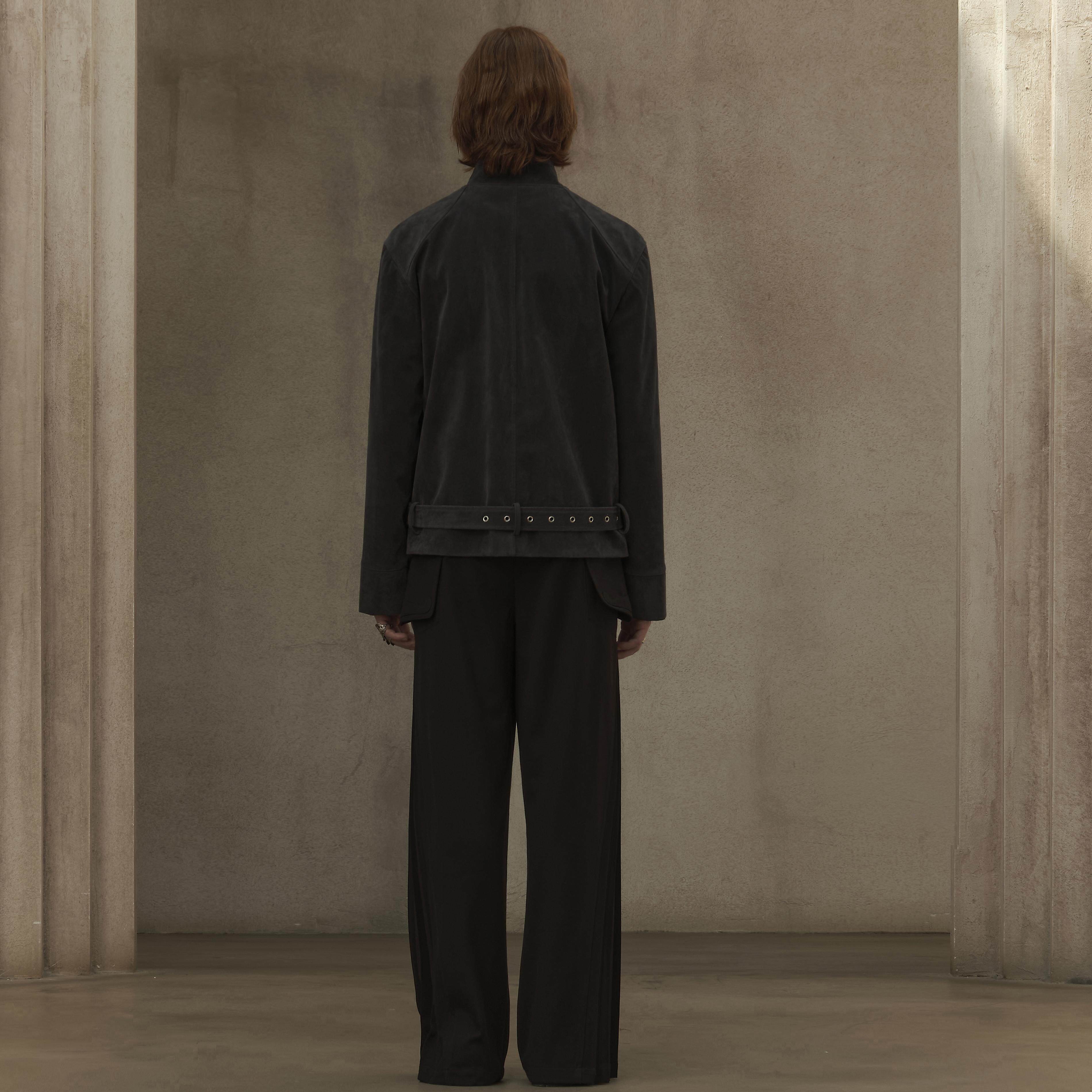 Person wearing a black suit standing in a minimalistic room with beige walls.