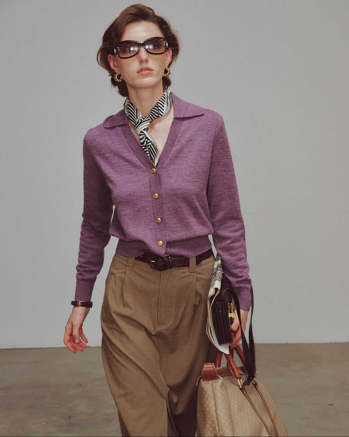 Woman wearing a purple cardigan and brown pants, holding a handbag against a plain background