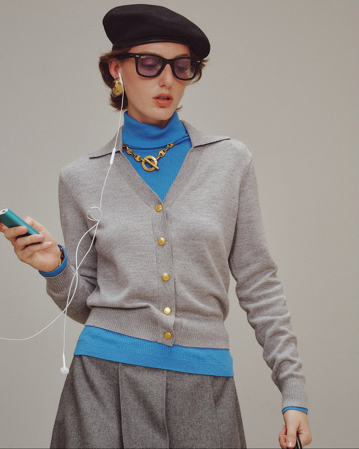 Woman wearing a gray cardigan, blue turtleneck, and sunglasses, holding a phone with earphones.