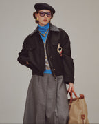 woman wearing a black jacket, gray pants, and holding a beige bag against a plain background