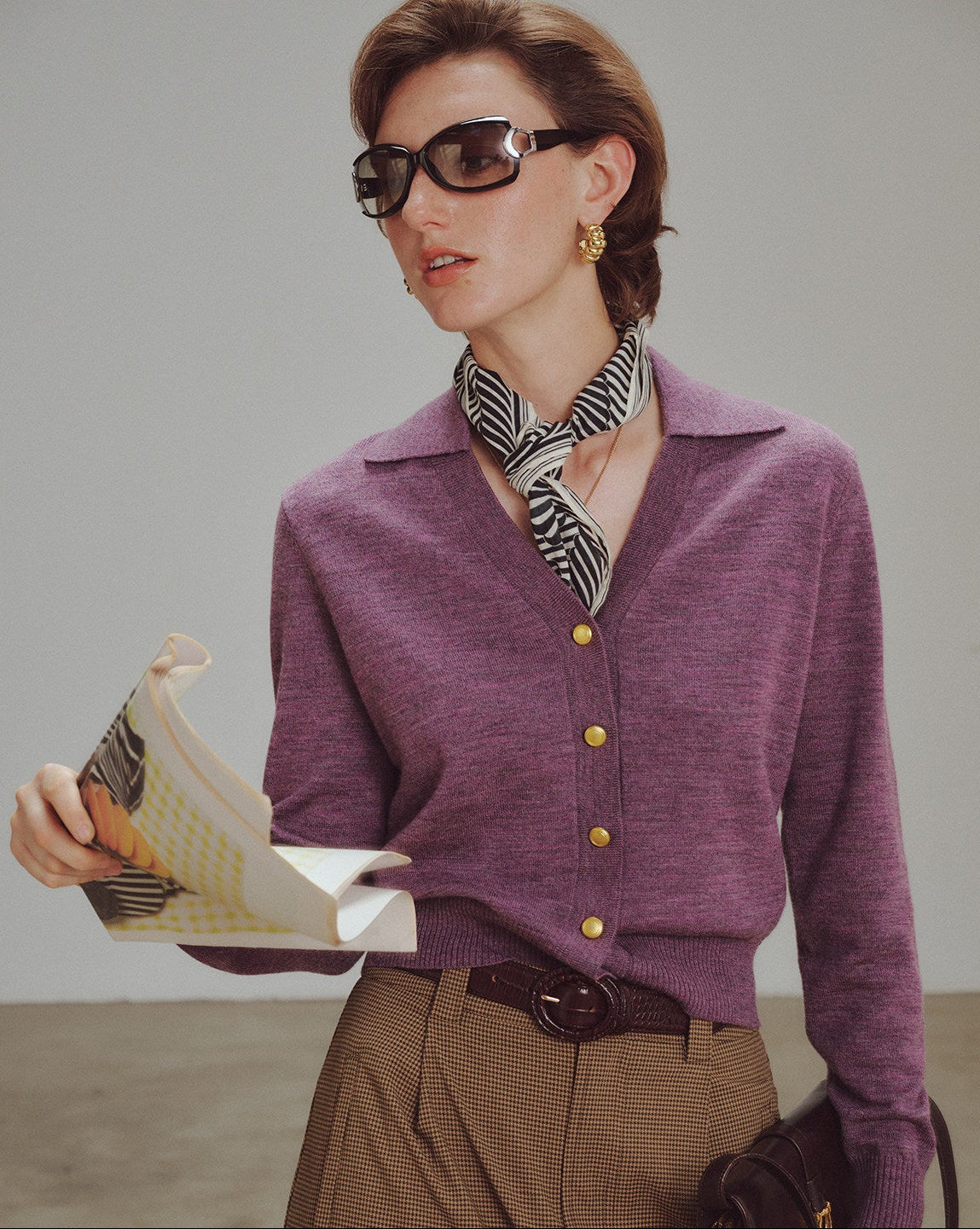 Woman wearing a purple cardigan with gold buttons, holding a book, and wearing sunglasses and a scarf.