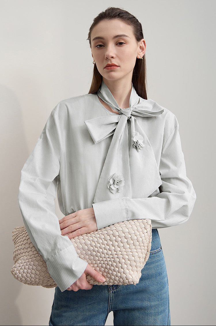 A woman models a light gray long-sleeve blouse with a large bow at the collar and decorative flower embellishments, paired with blue jeans and holding a beige woven clutch against a plain light gray background.