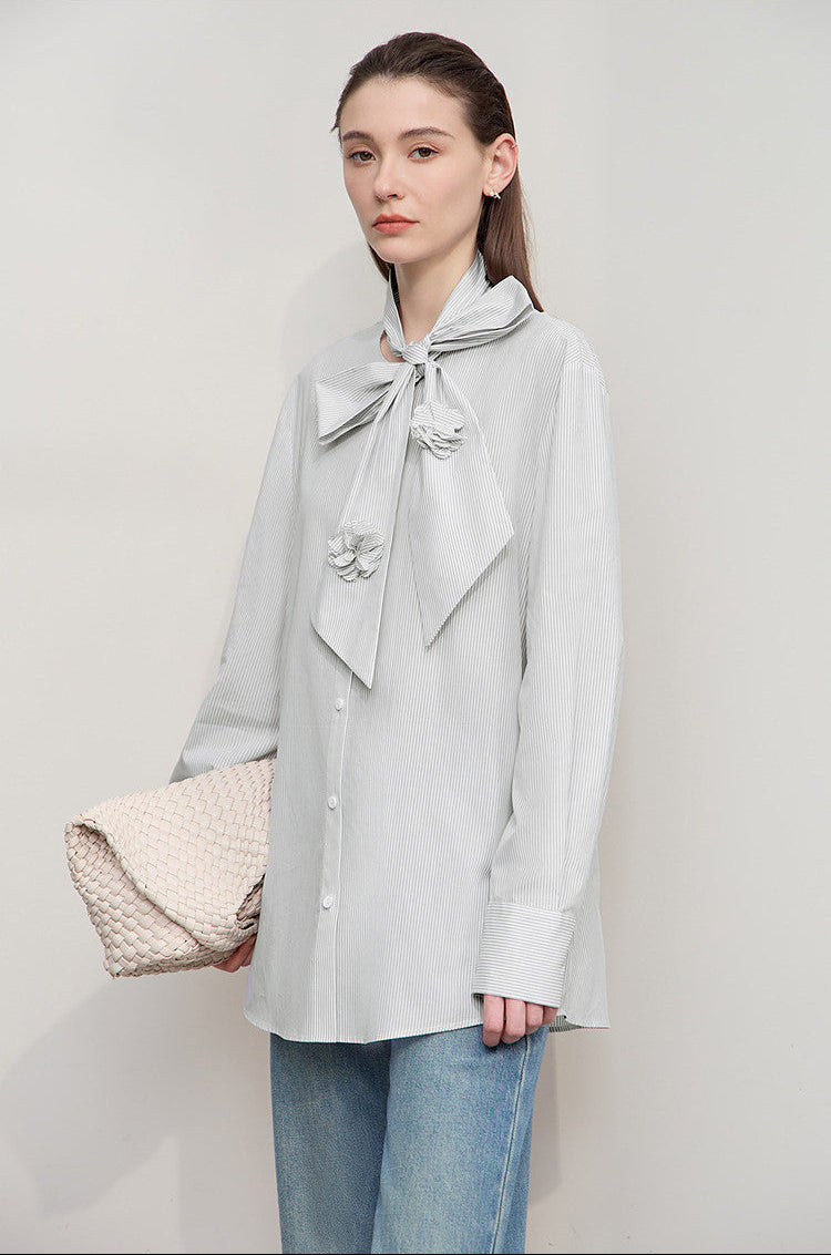 Woman wearing a light gray blouse with a large bow tie, holding a beige clutch against a plain background