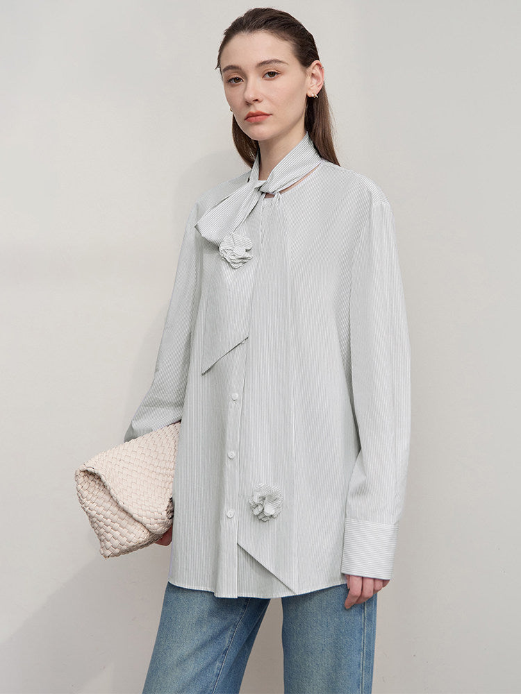 A woman modeling a light gray long-sleeve blouse with a large bow at the collar, decorative fabric flowers, and a button front, paired with blue jeans and holding a textured cream clutch against a plain light gray studio background.